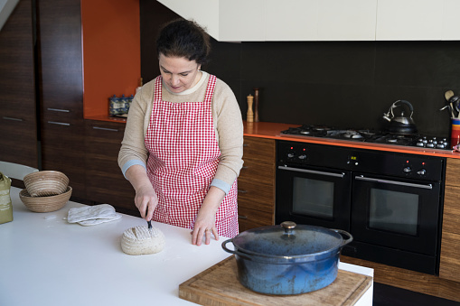 Partial front view of Caucasian woman wearing gingham apron and scoring top of dough before placing porridge loaf with oats and linseed in oven.