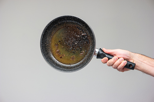 Caucasian male hand holding an old frying pan stained with brown burned oil and grease isolated on gray.