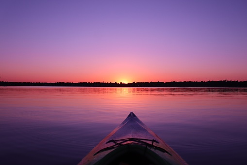 Kayak with bow turned towards the sun. Purple and pink vibrant colored sunset over reflective calm waters.