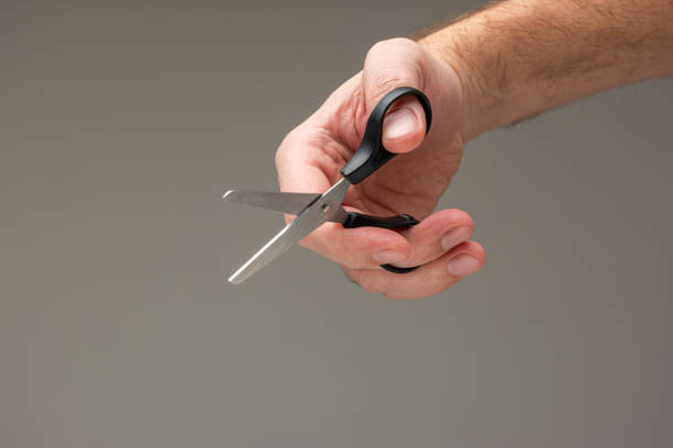 Caucasian male hand holding a small craft scissors with black plastic handles isolated on gray background.