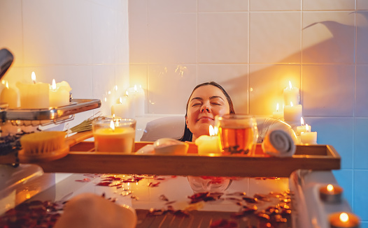 Young woman enjoying spiritual aura cleansing rose flower bath with rose petals and candles during full moon ritual. Body care and mental health routine.
