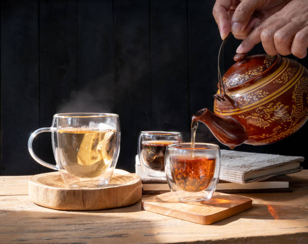 istock Hand pouring freshly brewed  tea glass cup with  traditional clay kettle and sugar cinnamon stick on dark wood table background 1299455776
