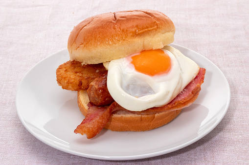 Breakfast brioche roll with a fried egg, hash brown, sausage, and bacon