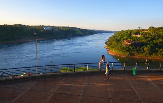 Puerto Iguazú, Argentina, November 18, 2019: Two girls take pictures at the confluence of the Iguazú and Paraná rivers on the tri-border at sunset. In the foreground is Argentina, on the left Paraguay and on the right Brazil.