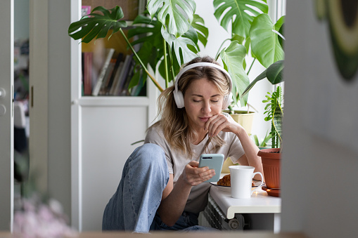 Mujer escuchando música usar auriculares blancos inalámbricos usando teléfono inteligente móvil, bebe té en casa photo