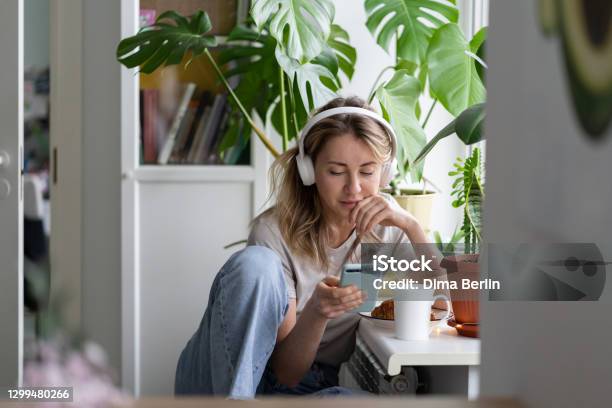 Woman Listening To Music Wear Wireless White Headphones Using Mobile Smart Phone Drinks Tea At Home Stock Photo - Download Image Now