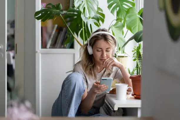 Woman listening to music, wear wireless white headphones, using mobile smart phone, chatting in social networks, sitting next to the window, houseplants on windowsill. Life at home. Time to relax.