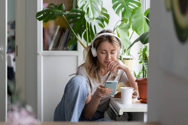 kobieta słuchając muzyki nosić bezprzewodowe białe słuchawki za pomocą telefonu komórkowego inteligentny telefon, pije herbatę w domu - słuchać zdjęcia i obrazy z banku zdjęć
