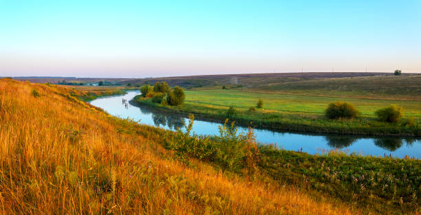 istock Sunny summer landscape with calm river flowing between the beautiful green hills,fields and meadows at quiet morning. 1299483195