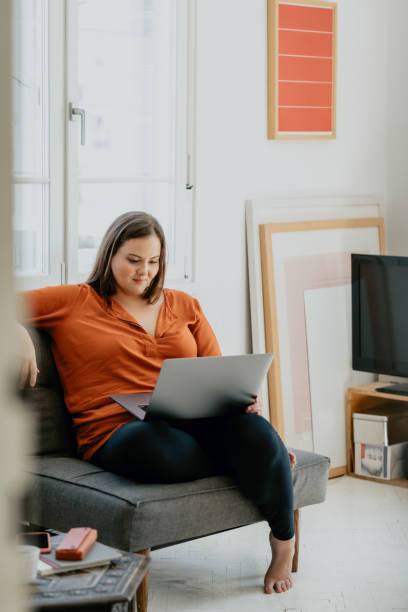 mujer de tamaño más sonriente sentada cómodamente en un sillón y trabajando en un ordenador portátil - living room elegance women long hair fotografías e imágenes de stock