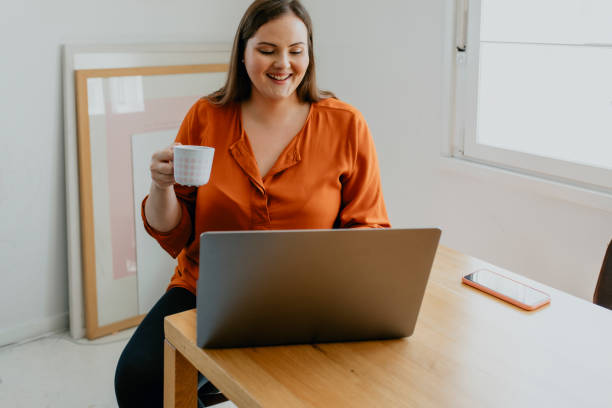 istock Happy  Plus Size Woman Enjoying a Cup of Coffee while Browsing the Web on her Laptop in the Morning 1299495979