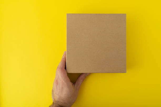 istock Square cardboard box in hand on yellow background. Copy space. Mock up. 1299496577