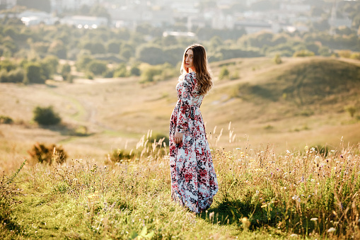 beautiful young woman in dress in floral print walking in the field at sunset. stylish romantic girl with long hair have a good time outdoors on city background. copy space.