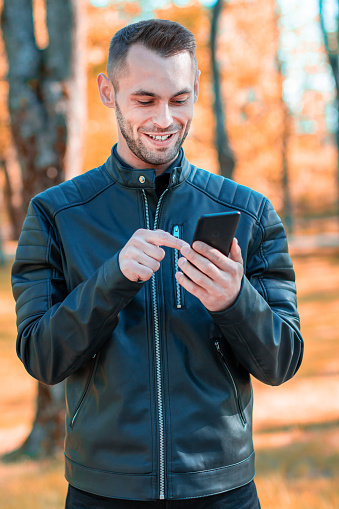 Youthful Satisfied Guy Using Black Smartphone at the Beautiful Autumn Park. Handsome Smiling Young Man with Mobile Phone at Sunny Day - Medium Shot Portrait