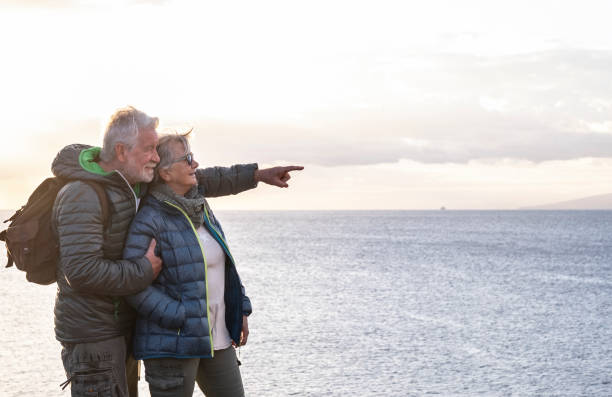 istock Two carefree senior people with backpack on their back enjoys the hike on the ocean cliffs looking at the horizon over water viewing the profile of an island and a ship. 1299503702