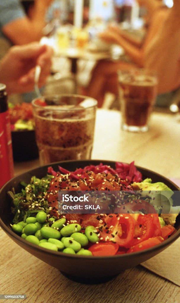 Delicious and fresh Hawaiin Poké Bowl A fresh and tasty poke bowl can be seen. It is topped with tuna, avocado, tomatoes, salad and edamame. In the background some non alcoholic beverages are being drunken. Bowl Stock Photo
