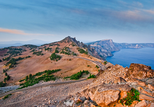 Hillman Peak is a basalt pinnacle on the rim of the Crater Lake caldera. At 8151 feet above sea level it is one of several peaks on the crater rim and the highest. This view of West Rim Drive, Llao Rock, Hillman Peak and Crater Lake was photographed at dusk from the Watchman Overlook in Crater Lake National Park, Oregon, USA.