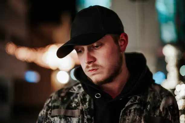 Bugged young man dressed with camouflage winter jacket and black baseball cap standing in illuminated city street at Night. Looking down to the fllor, with a irritated, bugged and pensive facial expression. Real People Millennial Generation Urban Night Portrait.