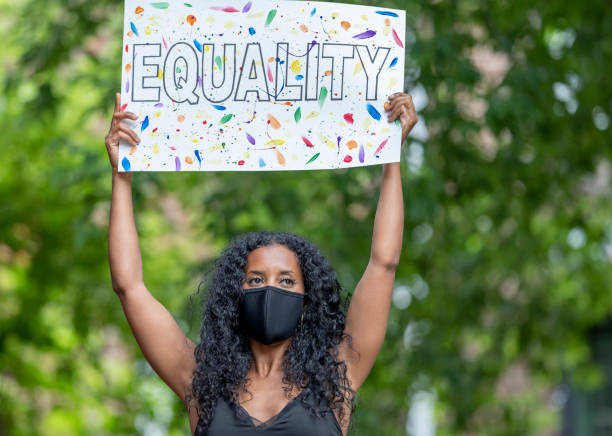 Equality for all A beautiful young female adult of African ethnicity is standing outdoors. She is wearing a face mask and is holding a poster with the word "Equality". civil rights stock pictures, royalty-free photos & images