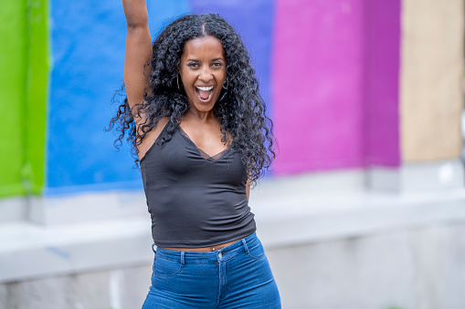 A beautiful young female adult of African ethnicity is standing in front of a pride mural. She has one hand up in the air and is smiling at the camera.