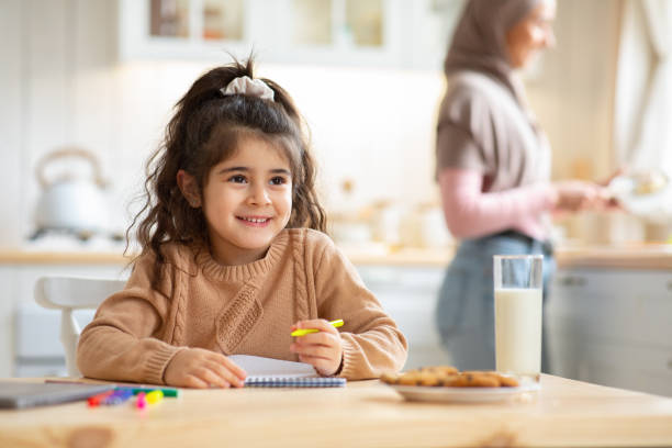 Cute Little Girl Drawing In Kitchen While Her Muslim Mom Cooking Lunch Cute Little Girl Drawing At Table In Kitchen While Her Muslim Mom In Hijab Cooking Lunch On Background. Adorable Female Child Using Colorful Pencils And Notepad, Enjoying Spending Time At Home arabic girl stock pictures, royalty-free photos & images