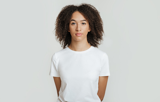 Serious, calm, strict emotions. Emotionless, unemotionally young african american woman with curly hair in white t-shirt, holds her hands behind back and looks in camera, isolated on white background