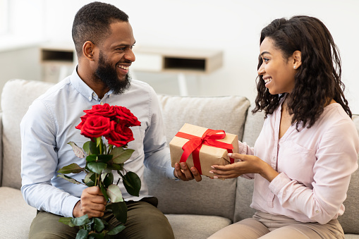 Romantic Gift. Black Man Holding Bunch Of Flowers And Giving Wrapped Box To His Woman Celebrating Holiday Together Sitting On Sofa At Home. Birthday Presents, Holidays Celebration Concept