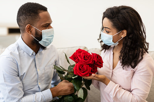 Dating During Pandemic. African american couple wearing medical face masks, spending time together at home, holding bouquet of red roses flowers. New normal, coronavirus, covid-19, quarantine