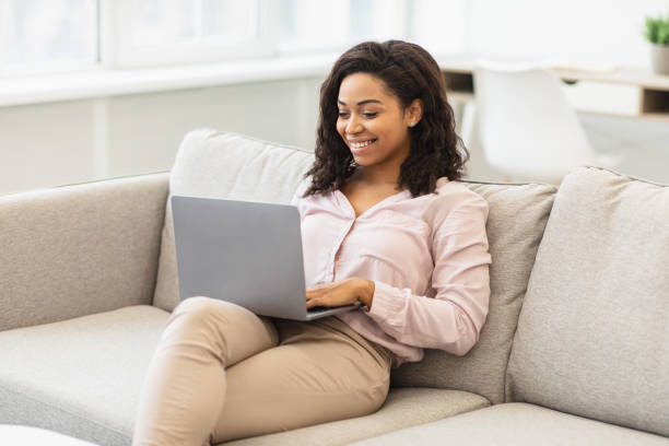 istock Black woman sitting on couch and working on laptop 1299555642