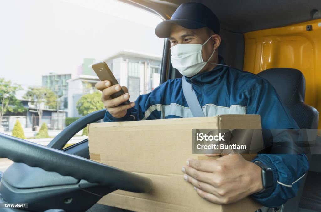Delivery man wif a parcel box Young Asian men wearing protective mask while working. delivery man and parcel box with smartphone in van.Professional truck driver, confident transportation service. Postman Check recipient address Truck Driver Stock Photo