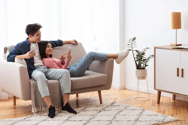 Japanese Couple Using Cellphones Texting And Browsing Internet Relaxing Sitting On Couch Indoor. Weekend Leisure. Happy And Relaxed Asian Family Using Mobile Application Resting At Home