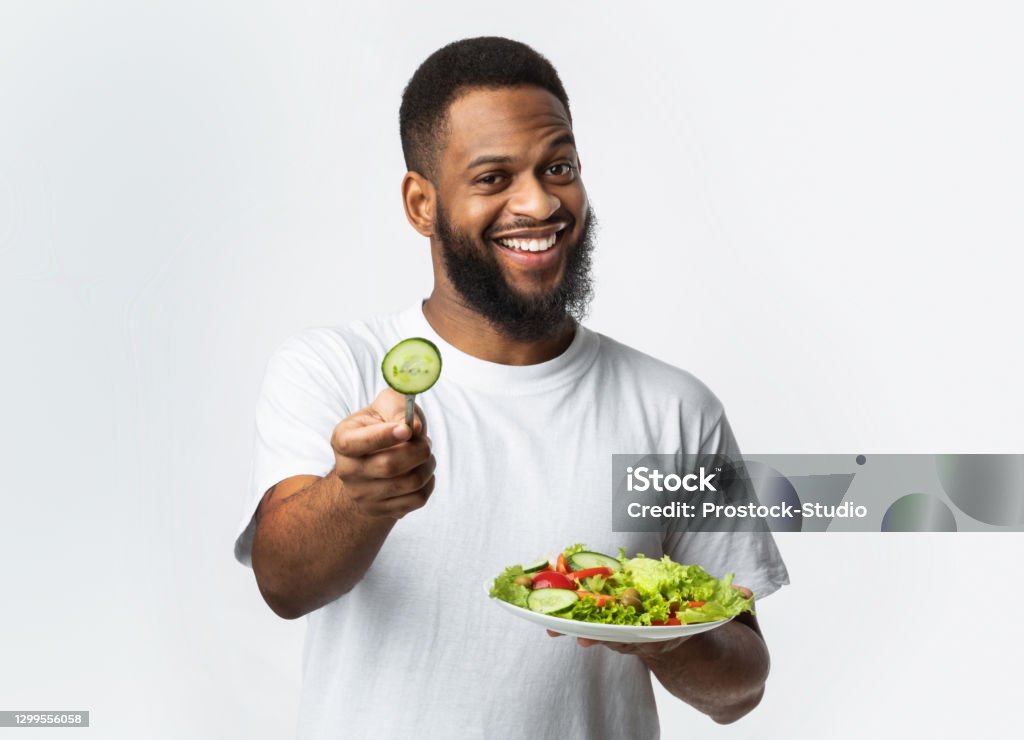 Cheerful Black Man Holding Salad Standing On White Background, Studio Cheerful Black Man Holding Salad Offering Slice Of Cucumber To Camera Recommending Healthy Eating Standing On White Background. Studio Shot. Try Vegan Diet Concept Healthy Eating Stock Photo