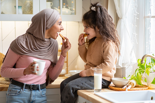 Muslim Mom In Hijab And Her Little Daughter Eating Snacks In Kitchen, Having Fun Together At Home. Happy Islamic Family Mother And Child Enjoying Milk And Homemade Cookies, Free Space