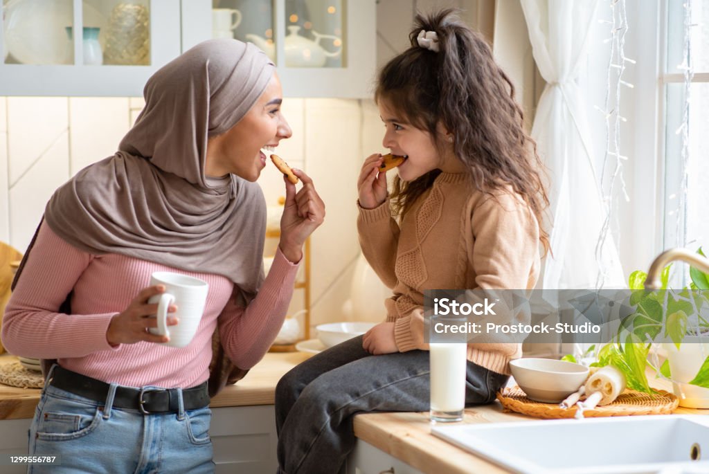 Muslim Mom In Hijab And Her Little Daughter Eating Snacks In Kitchen Muslim Mom In Hijab And Her Little Daughter Eating Snacks In Kitchen, Having Fun Together At Home. Happy Islamic Family Mother And Child Enjoying Milk And Homemade Cookies, Free Space Child Stock Photo