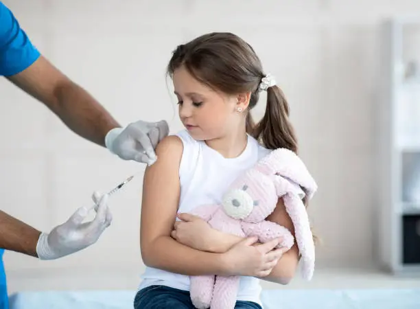 Doctor doing injection of coronavirus vaccine to little girl with toy bunny at clinic. Cute child on visit to medical specialist, getting immunization against coronavirus. Medicine and vaccination