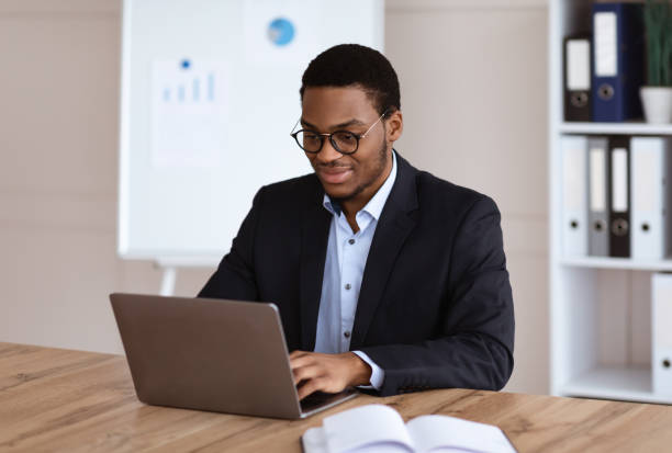 Young black entrepreneur working with laptop, office interior Smiling young black entrepreneur in formal suit working with laptop, wearing glasses, office interior, copy space. Successful millennial african american businessman working on business project rich black men pictures stock pictures, royalty-free photos & images