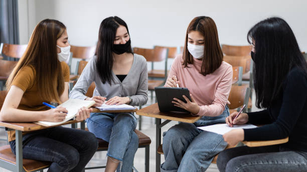 istock Group of diverse international students wearing protective  masks and talking, discussing the project, sitting at the desk in the classroom at the university 1299559689
