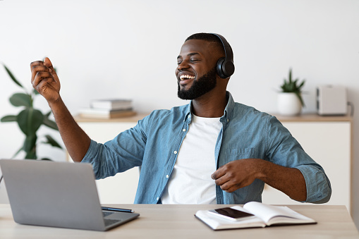 Work With Fun. Cheerful Black Freelancer Man In Wireless Headphones Listening Music And Playing Virtual Guitar At Workplace, Resting At Desk In Home Office, Enjoying Remote Job Opportunities