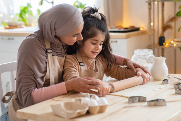 милая маленькая девочка и мусульманская мама свертывая тесто вместе на кухне - свертывая стоковые фото и изображения