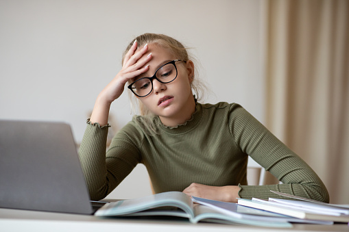 Teenager blonde girl in glasses having difficulties with studying from home, sitting at desk with laptop and books, touching her had, copy space. Upset school girl got tired from studying