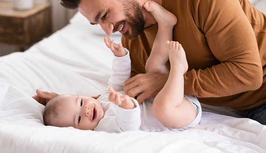 Happy Father And Little Baby Playing Together Having Fun Bonding Lying On Bed At Home. Young Dad Enjoying Parenting Cuddling With Toddler Child. Fatherhood Joy Concept. Cropped