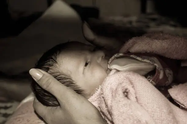 Photo of Beautiful Newborn baby boy closeup (6 days old) lying in mother lap in prenatal hospital. Kid wrapped in baby blanket (warm clothing) smiling and looking. His mother consoling and resting her baby.