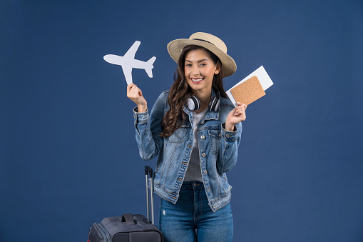 Happy young Asian woman holding passport with boarding pass and airplane mockkup white paper on blue color background, holiday and travel concept