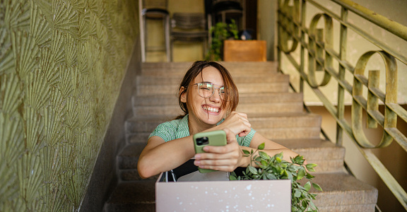Photo of a young woman sitting on the stairs and checking her phone while moving into the new building; checking if her furniture is on the way to her new home address.