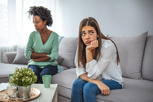 Two upset friends not talking to each other after fight on the sofa in sitting room at home. Two women friend sulking at each other, bad relationship concept.  Friendship, quarrel, female disagreement, copy space