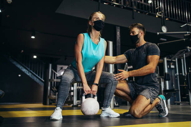 Sporty people exercising in fitness gym Young fit and attractive woman at body workout in modern gym together with her personal fitness instructor or coach. They keeping distance and wearing protective face masks. Coronavirus world pandemic and sport theme. fitness trainer stock pictures, royalty-free photos & images