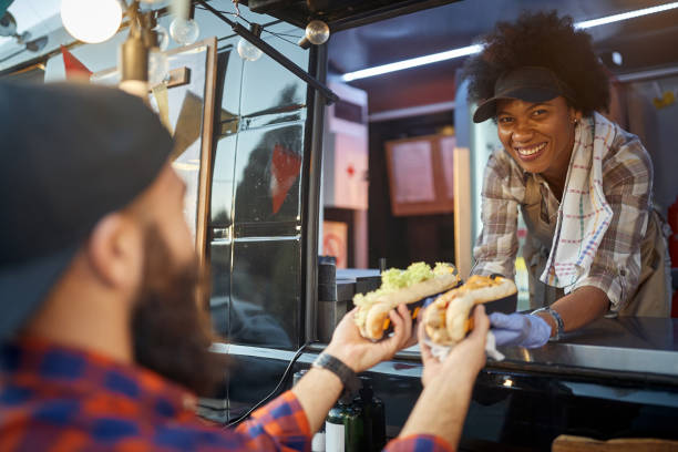 istock beautiful young afro-american female employee looking at camera, smiling, while giving sandwiches through a fast food window to a caucasian male customer 1299587888