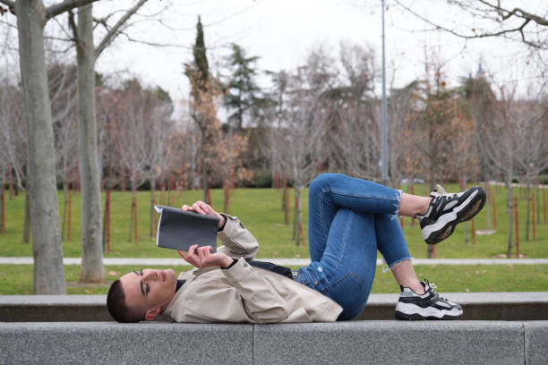 istock Handsome young man wearing make up, reading a book lying on a wall with his feet up. Non binary androgynous guy. 1299603985