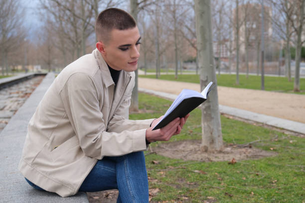 istock Handsome young man wearing make up, reading a book sitting on a wall. Non binary androgynous guy. 1299604238