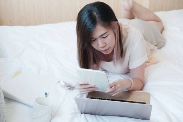 mulher jovem asiática sorridente está deitada em cama branca segurando tablet para trabalho online e compras com laptop em medea social no quarto. empresária trabalha em casa com conceito de tecnologia. - business mobile phone telephone women - fotografias e filmes do acervo